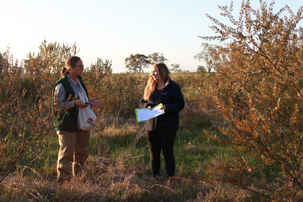 Two women, one with a bag and the other with a clipboard standing among native shrubs.